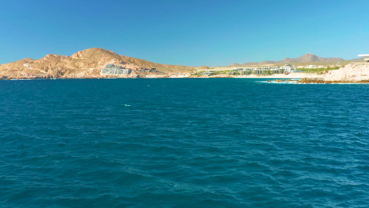 Opening view over turquoise water toward Chileno Beach Los Cabos, with distant shoreline buildings, tan mountains, clear blue sky, and no visible people.