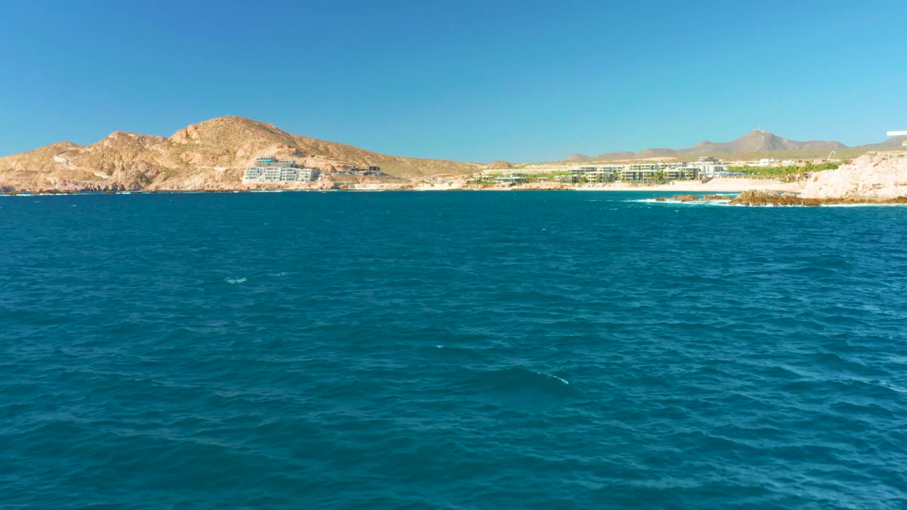Calm blue sea with a pale rocky shore, beachfront buildings, and mountains beyond.