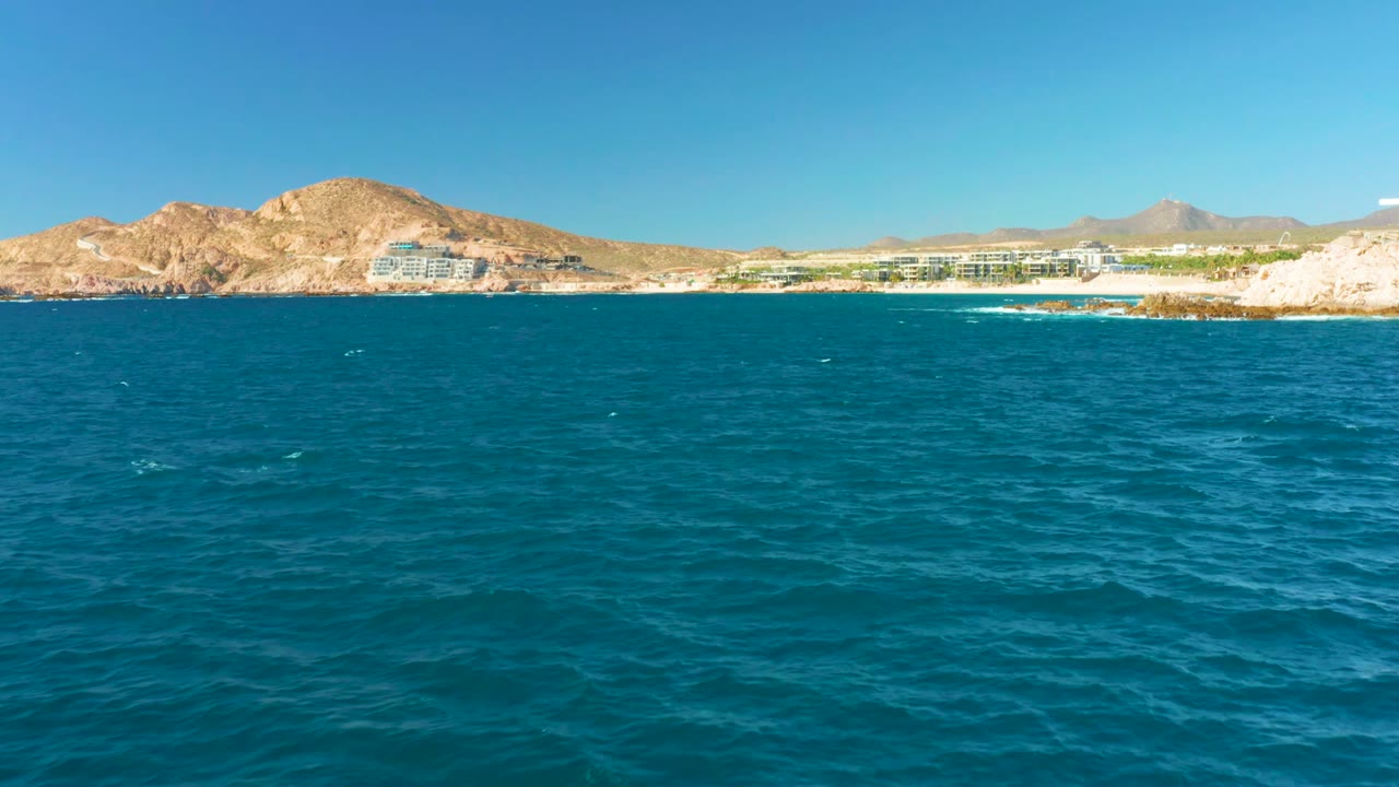 A broad foreground of textured ocean leads to distant Chileno Beach buildings and desert-colored mountains under a clear blue sky, with no visible people.