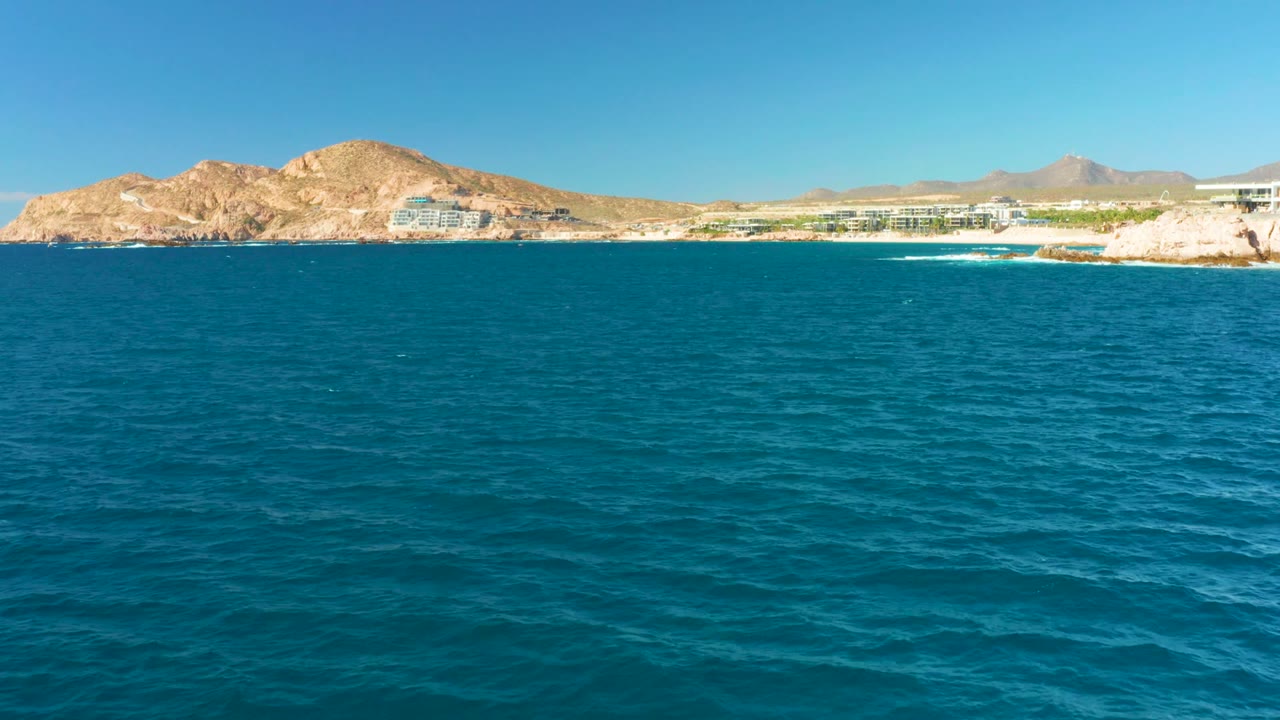 Wide view of blue water at Chileno Beach Los Cabos with tan rocky hills, low shoreline buildings, clear sky, and no visible people at the start of the clip.