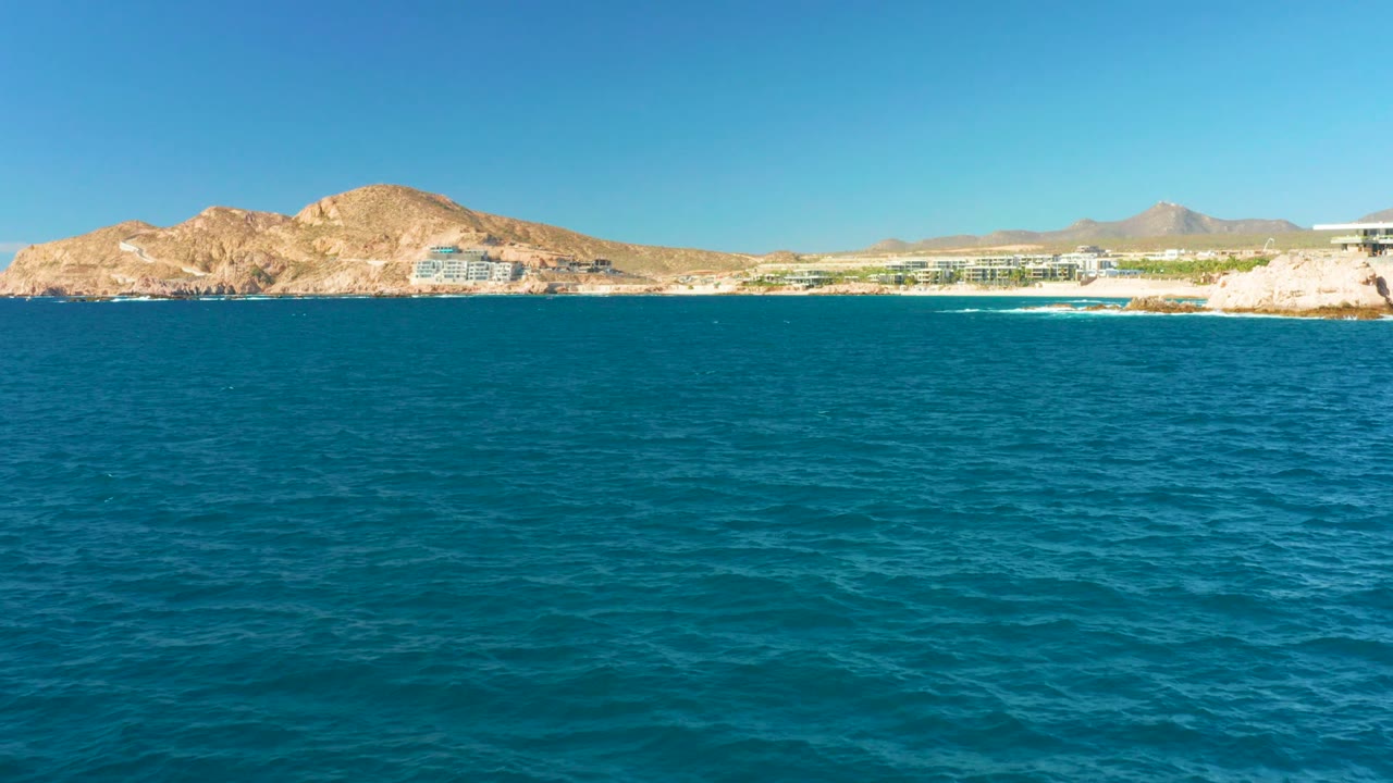 A broad band of blue sea faces the Chileno Beach Los Cabos coast, with tan rocky hills, low buildings along shore, a pale headland, and no visible people.