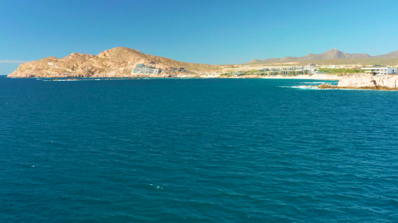 At 0:00, a wide aerial view at Chileno Beach Los Cabos shows deep blue ocean in the foreground, rocky shoreline, low coastal buildings, tan hills, distant mountains, clear sky, and no visible people.