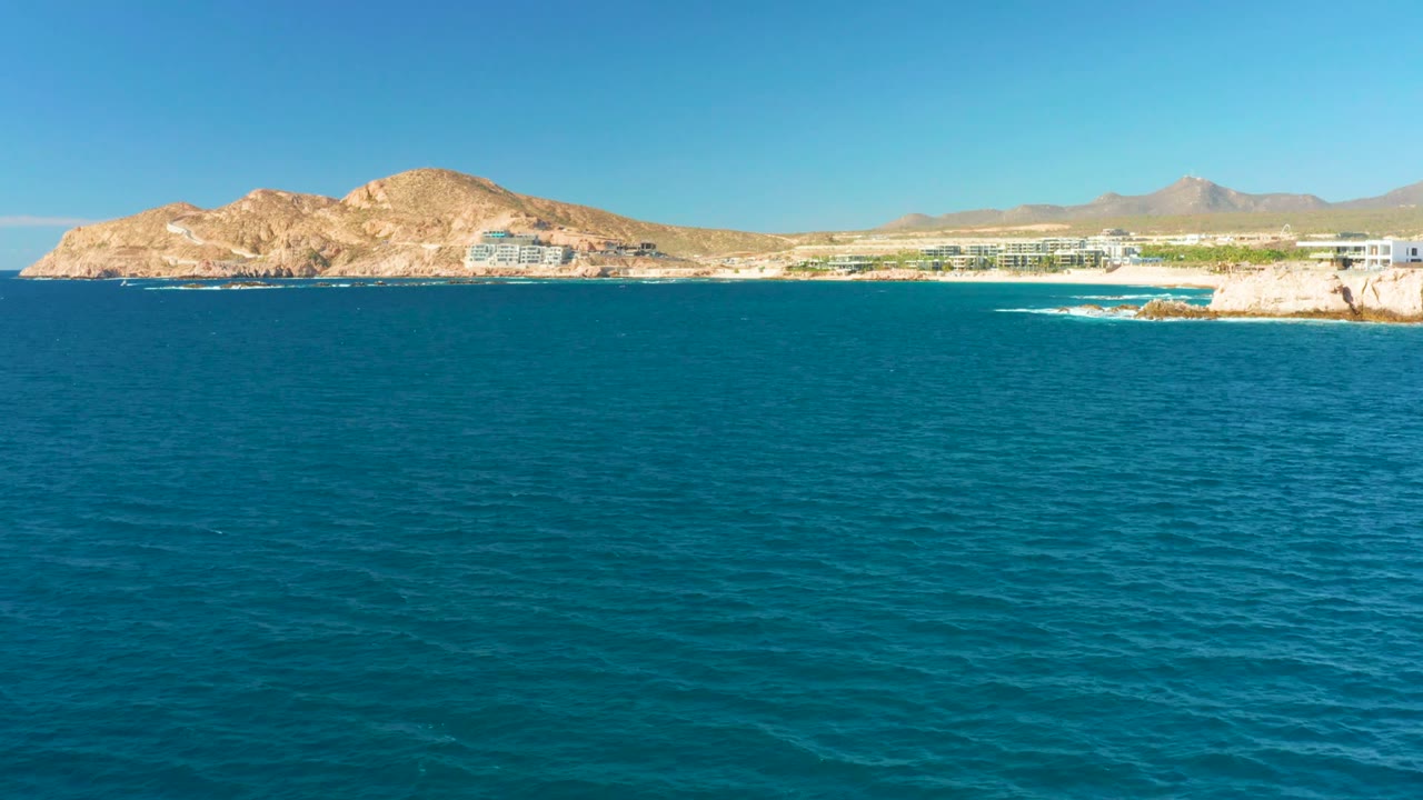 At 0:01, the ocean surface fills the lower frame while the Chileno Beach Los Cabos coastline runs across the horizon with pale buildings, tan rock, distant mountains, and no visible people.