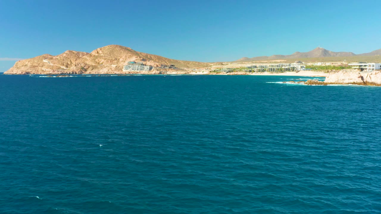 At 0:02, the Chileno Beach Los Cabos shore remains distant behind broad blue water, with a rocky point on the left, light buildings near the coast, mountains in the background, and no visible people.