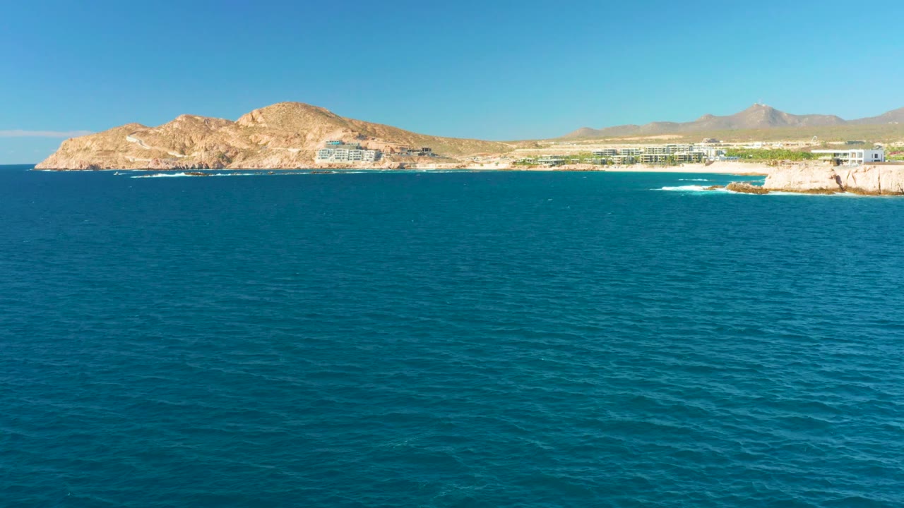 Opening frame of Chileno Beach in Los Cabos with turquoise ocean in the foreground, tan hills and coastal buildings on the horizon, a small white wave near the right shore, and no visible people.