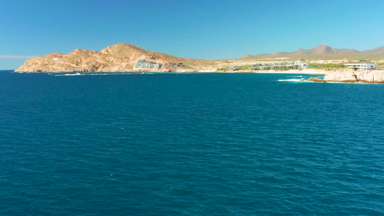 Calm blue ocean facing a rocky shoreline with low buildings and hills beyond.