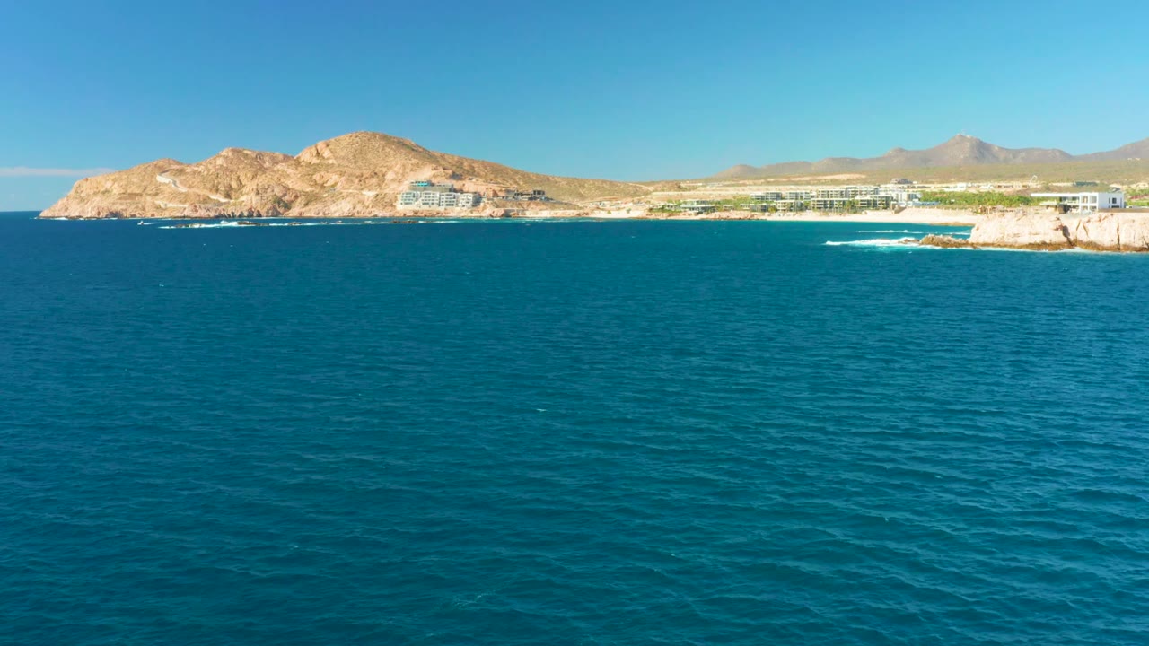 At 0:02, turquoise water dominates the frame while Chileno Beach shoreline buildings sit under dry hills, and a narrow white wave breaks against the rocky point on the right with no visible people.