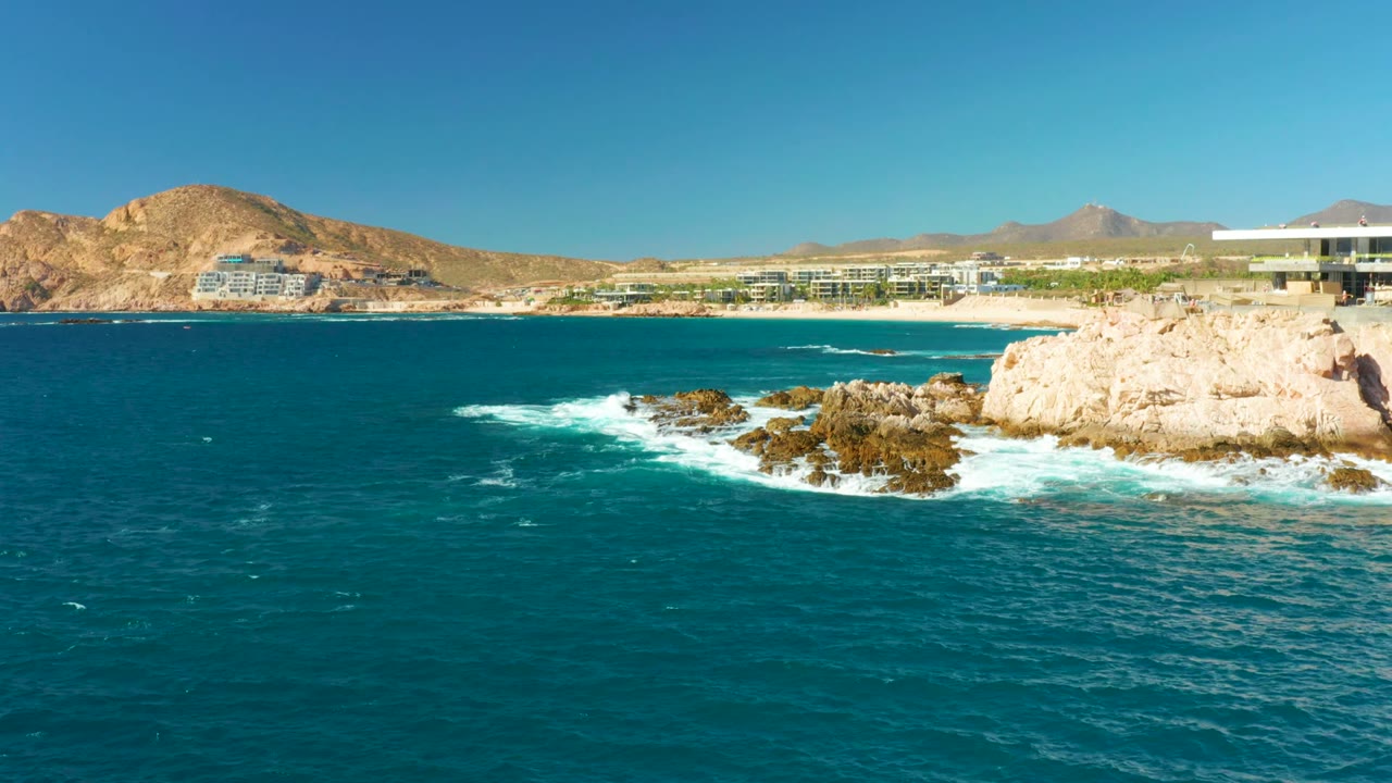 Wide elevated view at Chileno Beach Los Cabos with turquoise ocean in front, white surf around dark rocks, pale cliffs, coastal buildings, desert hills, and no visible people.