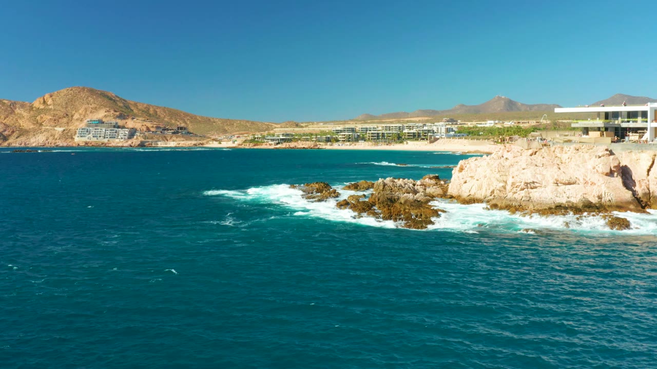 Ocean waves spread white foam around rocks below coastal buildings.