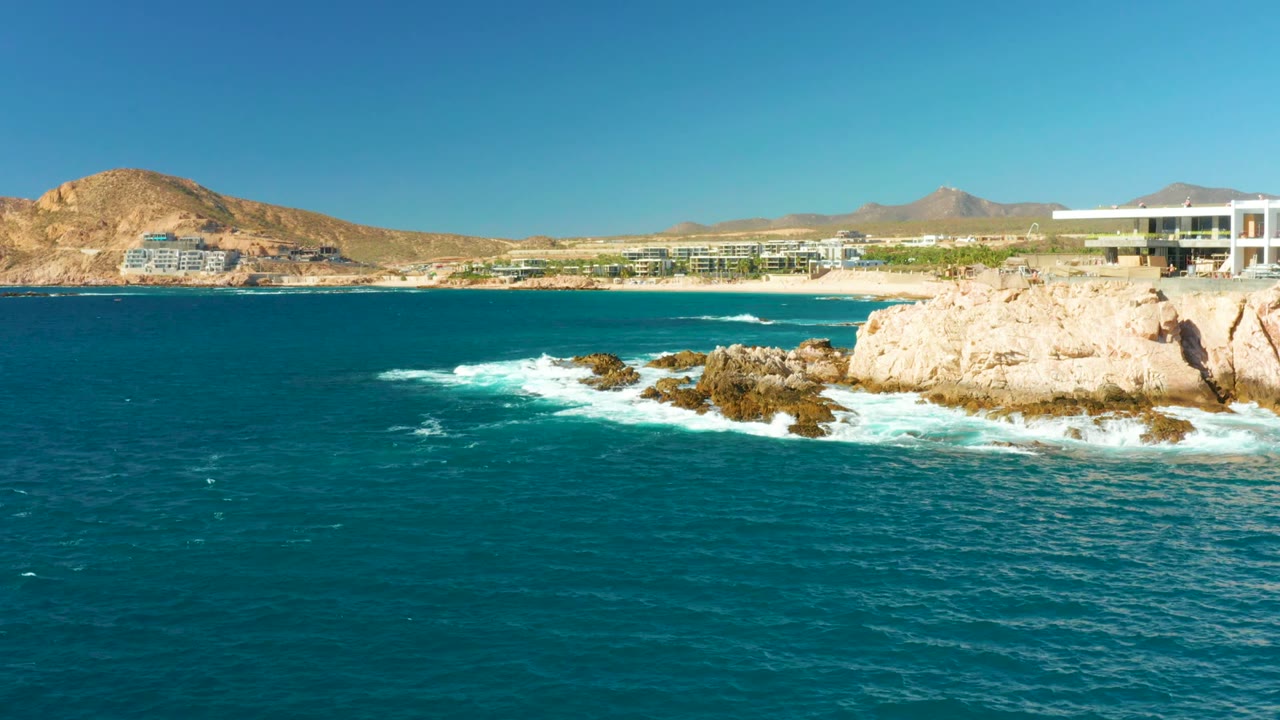 At 0:02, turquoise water fills most of the frame while surf wraps the central rocks, pale cliffs sit to the right, coastal buildings run along the beach, and no visible people appear.