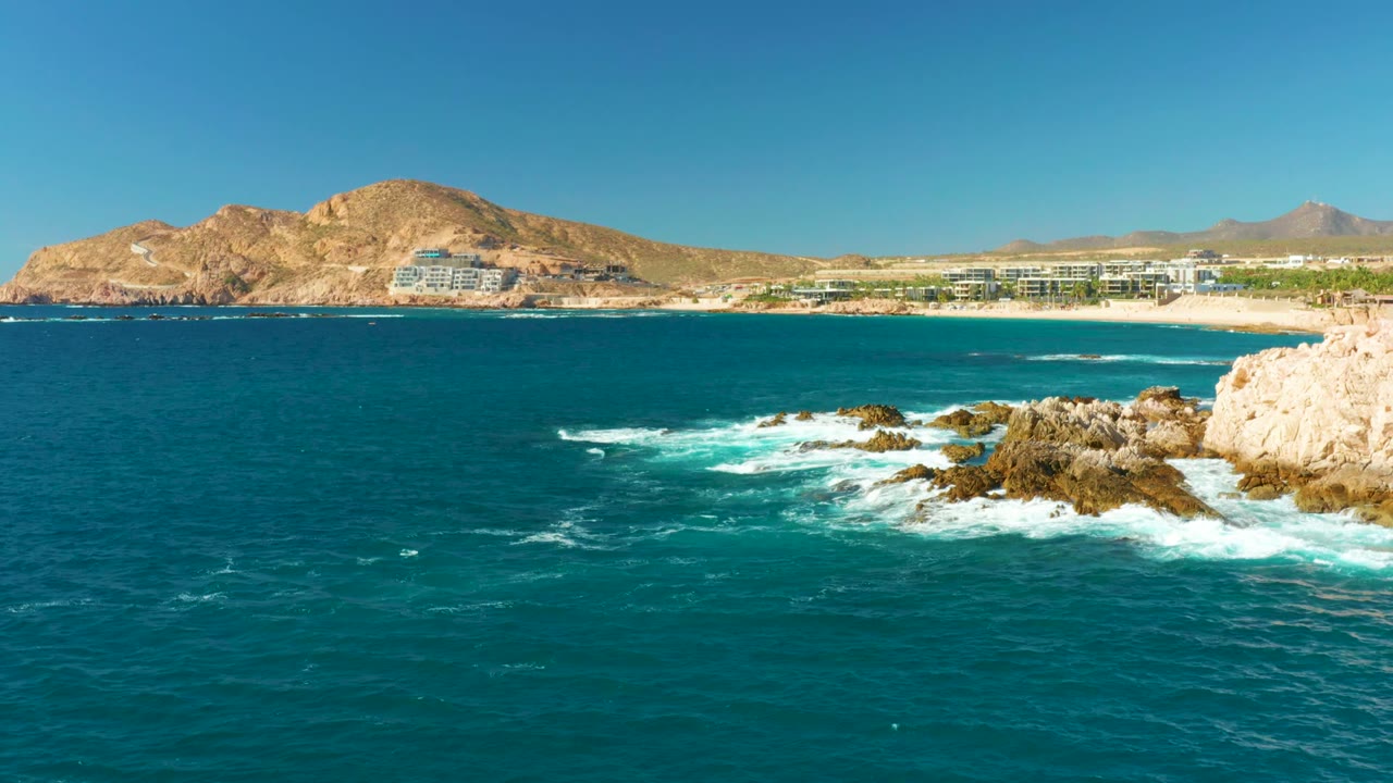 At Chileno Beach in Los Cabos, deep blue water fills the foreground while white surf breaks on dark rocks, with beach, resort buildings, dry mountains, clear sky, and no visible people.