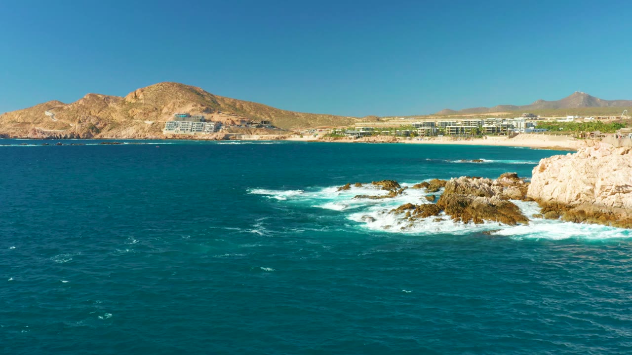 At 0:02, a bright section of pale cliff enters at right while waves wrap the rocks, backed by Chileno Beach, resort structures, dry mountains, and no visible people.