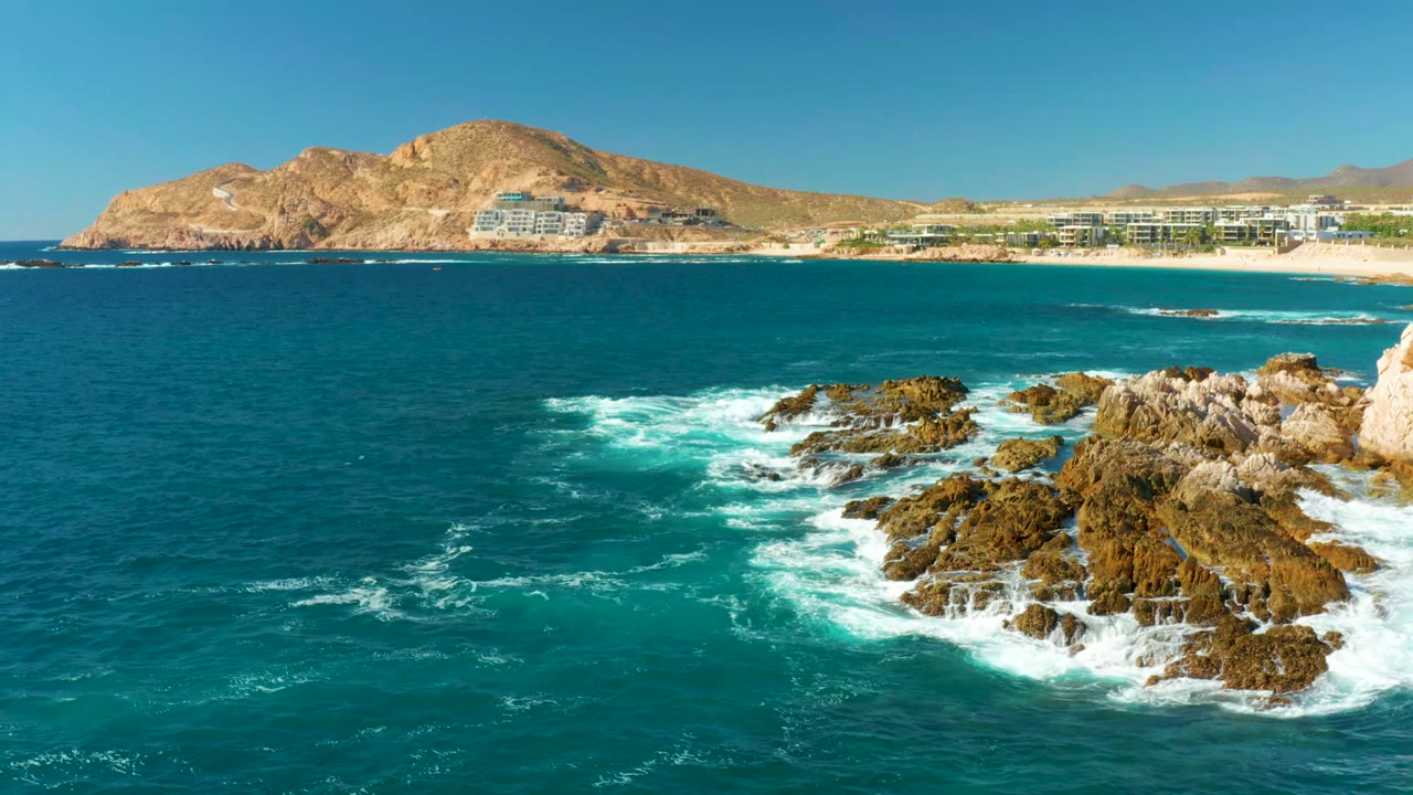 At Chileno Beach Los Cabos, turquoise water and white surf surround dark rocky outcrops, with beach resorts and tan mountains in the background and no visible people.