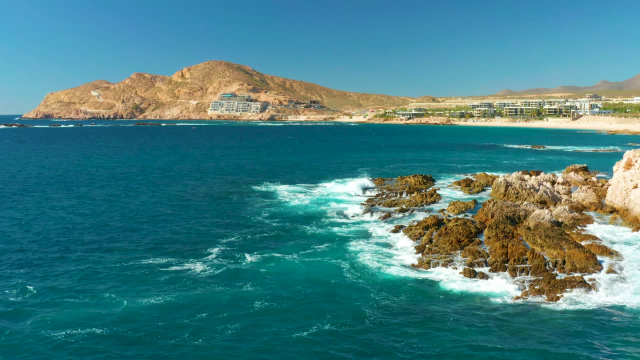 White waves spreading over rocks with open blue water, beach buildings, and mountains beyond.