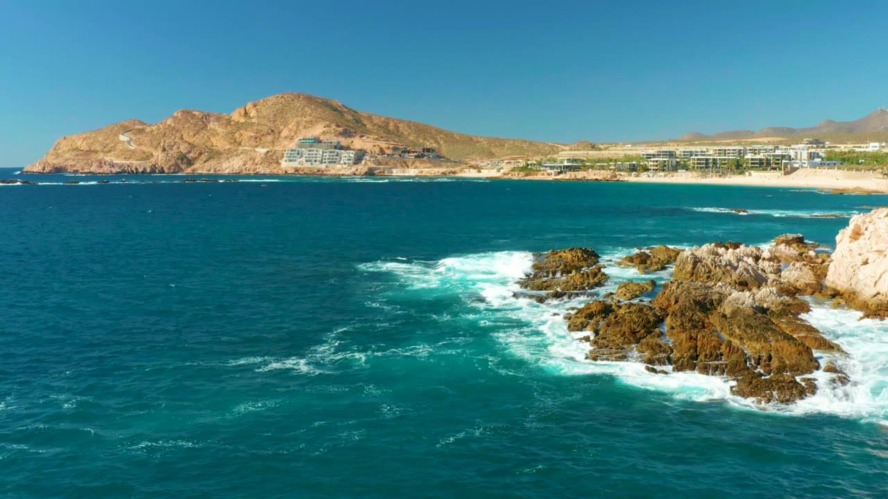 At Chileno Beach Los Cabos, the rocks sit slightly right of center as foamy surf curls around them, backed by beach resort buildings, arid mountains, and no visible people.