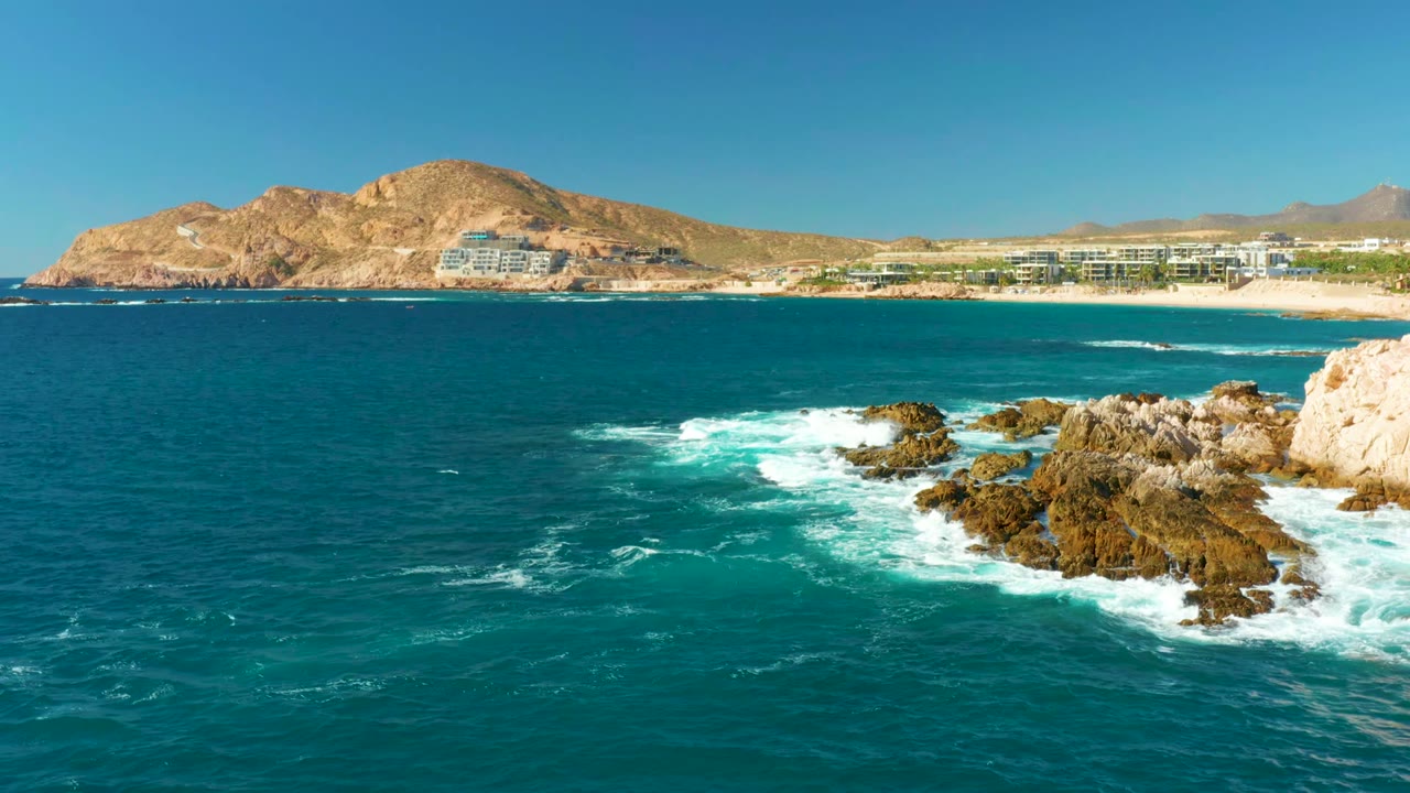 Ocean surf crashes across rocks near a pale cliff with beach resorts and mountains in background.