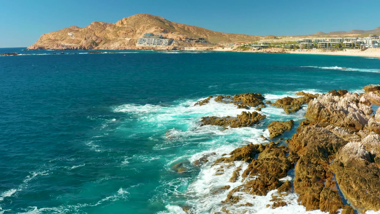 At Chileno Beach in Los Cabos, turquoise water breaks over jagged foreground rocks while a sandy beach, resort buildings, dry hill, and clear sky sit in the distance, with no visible people.