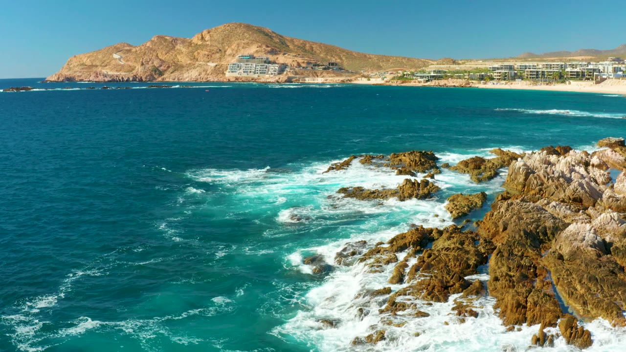 White surf spreading around rocks below a beach and hillside buildings