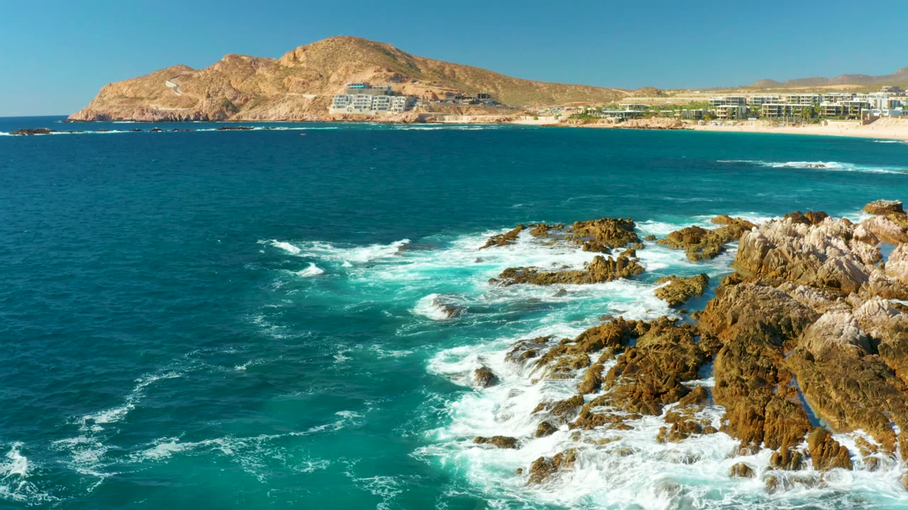 At Chileno Beach in Los Cabos, fresh white surf hits the right-side rocks and curls through turquoise water, with the distant sand, beachfront buildings, tan hill, and cloudless sky visible, with no visible people.