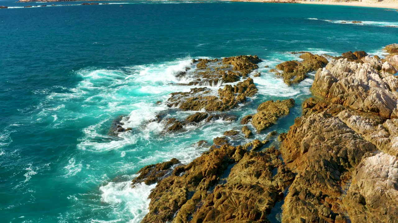 Aerial view of turquoise surf breaking around jagged rocks at Chileno Beach in Los Cabos, with pale cliffs at right and no visible people.