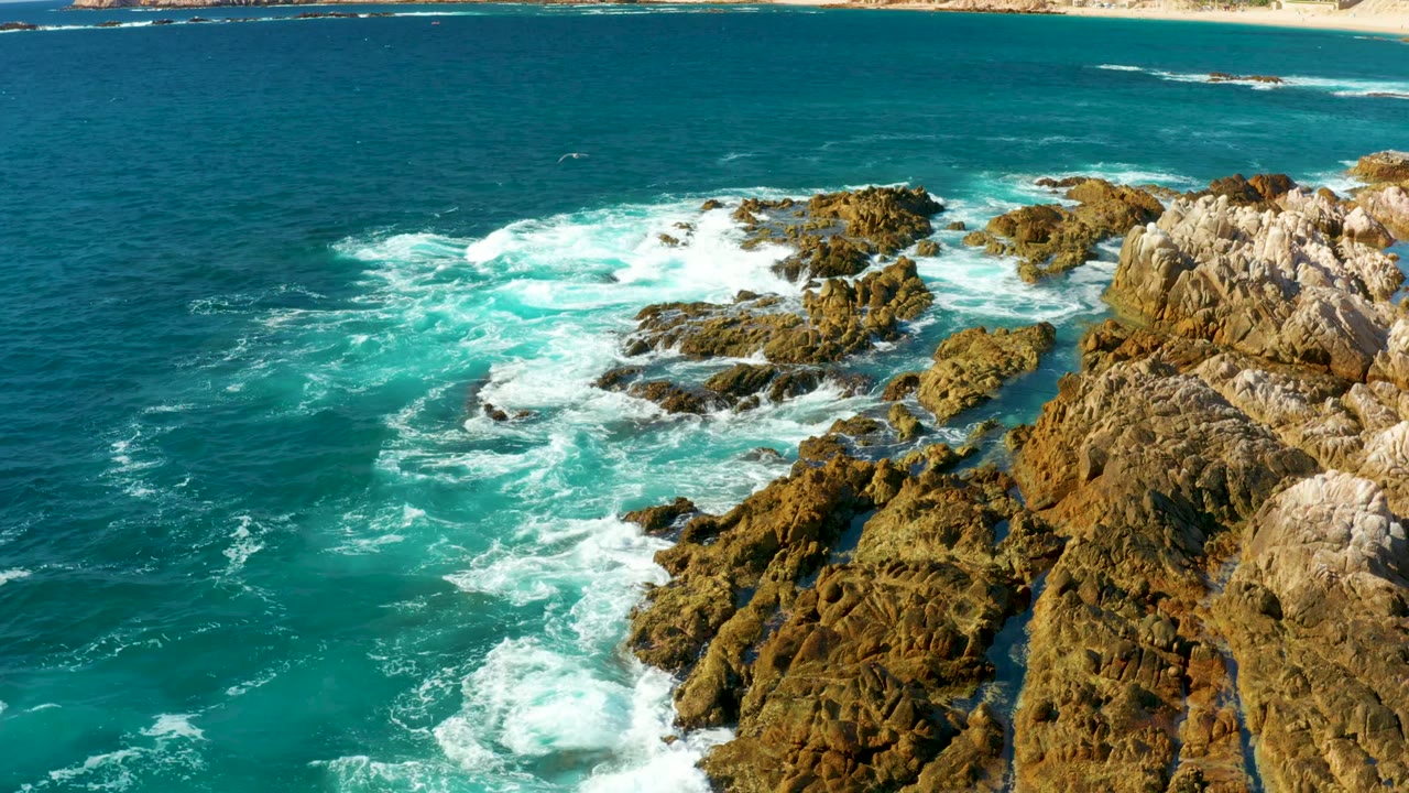 Aerial coastal frame at Chileno Beach showing waves washing around clustered rocks, a bright teal foam patch, and pale cliff faces at right, with no visible people.