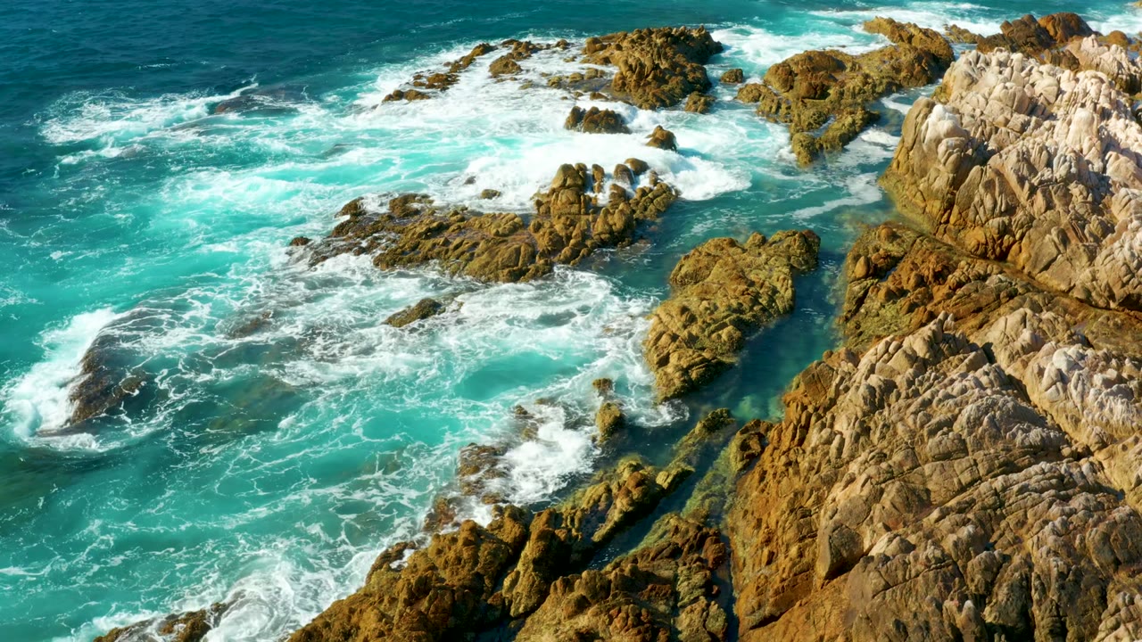 Aerial view of Chileno Beach Los Cabos surf washing through tan rock formations, with bright turquoise water and no visible people.