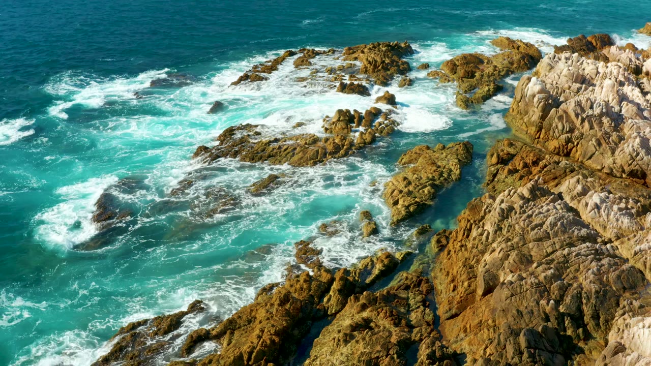 Aerial view of Chileno Beach Los Cabos waves breaking over a broad rocky shelf, with open teal water above and no visible people.