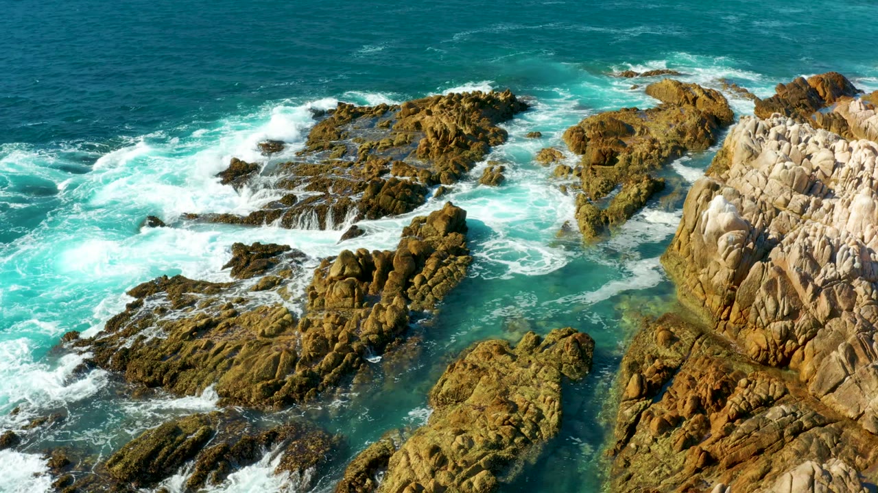 Aerial frame at Chileno Beach in Los Cabos showing turquoise water and white foam wrapping around brown rock outcrops, with no visible people.
