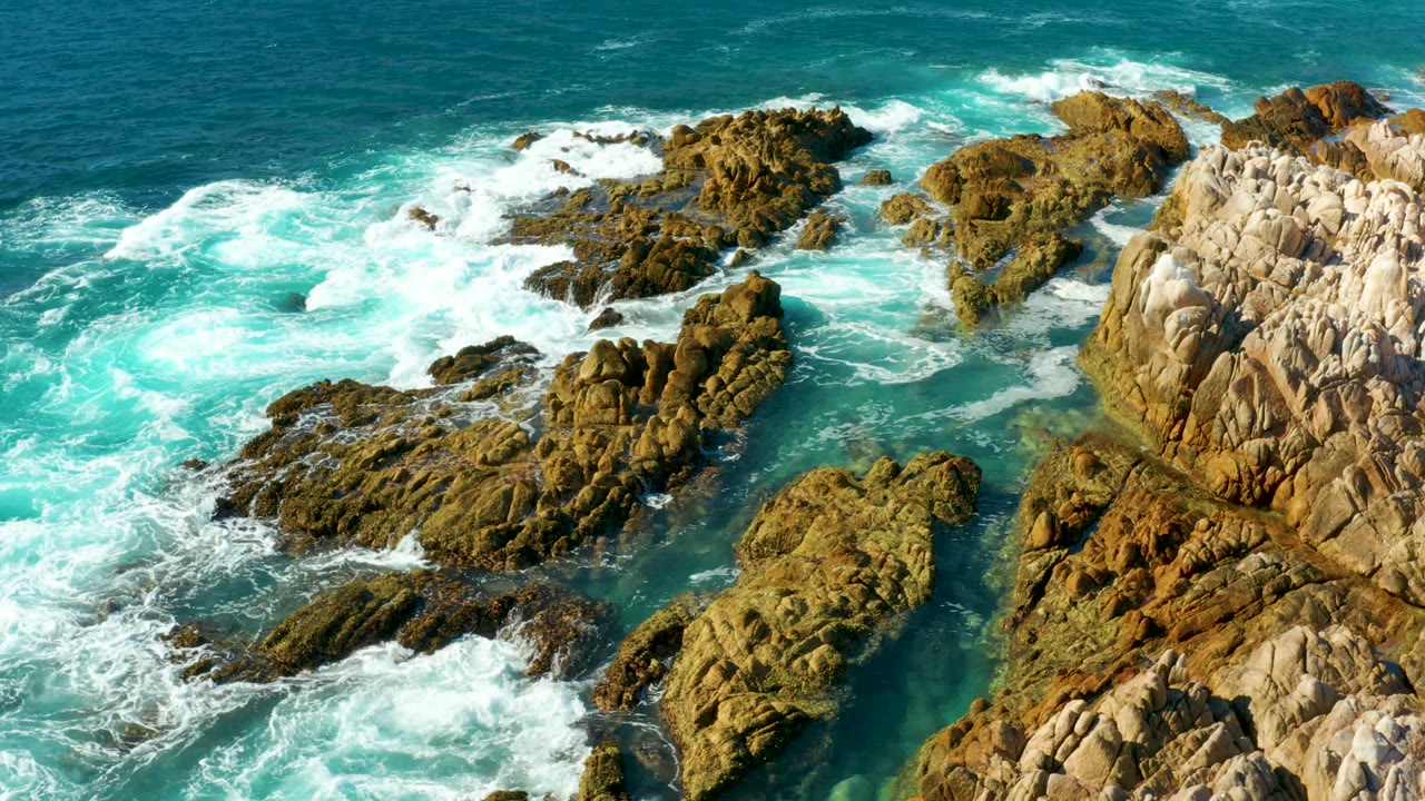 Chileno Beach Los Cabos aerial frame showing breaking waves over clustered brown rocks, teal water pools, and pale coastal stone on the right, with no visible people.