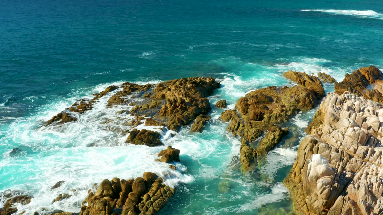 Overhead view at Chileno Beach in Los Cabos showing turquoise surf wrapping around tan and dark rock formations, with open ocean beyond and no visible people.