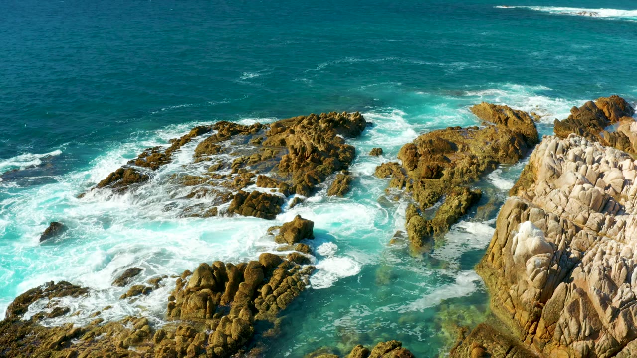 High drone view at Chileno Beach with foamy water pushing between clustered brown rocks and the larger pale rock wall at right, with no visible people.
