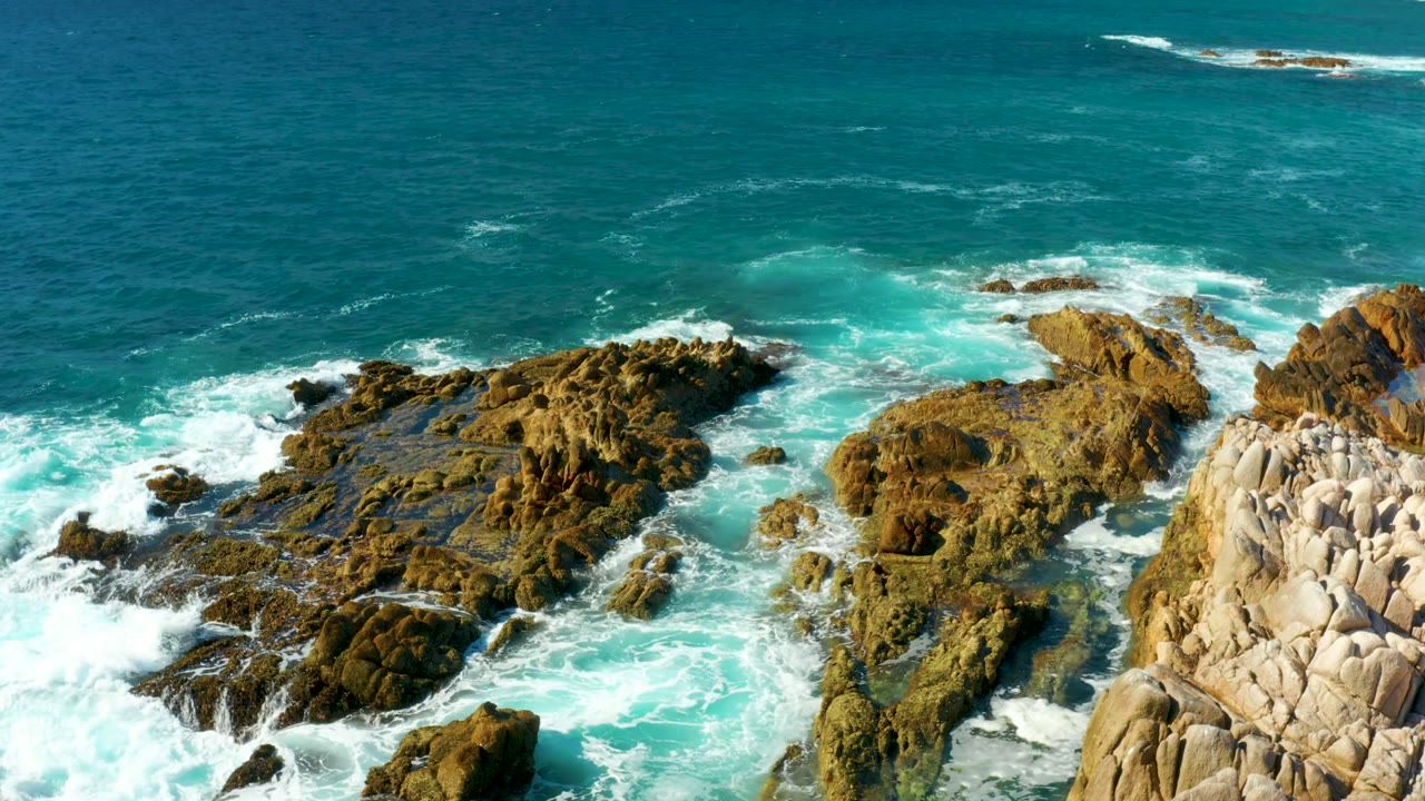 Aerial view of turquoise ocean surf breaking around brown rock shelves at Chileno Beach in Los Cabos, with no visible people.