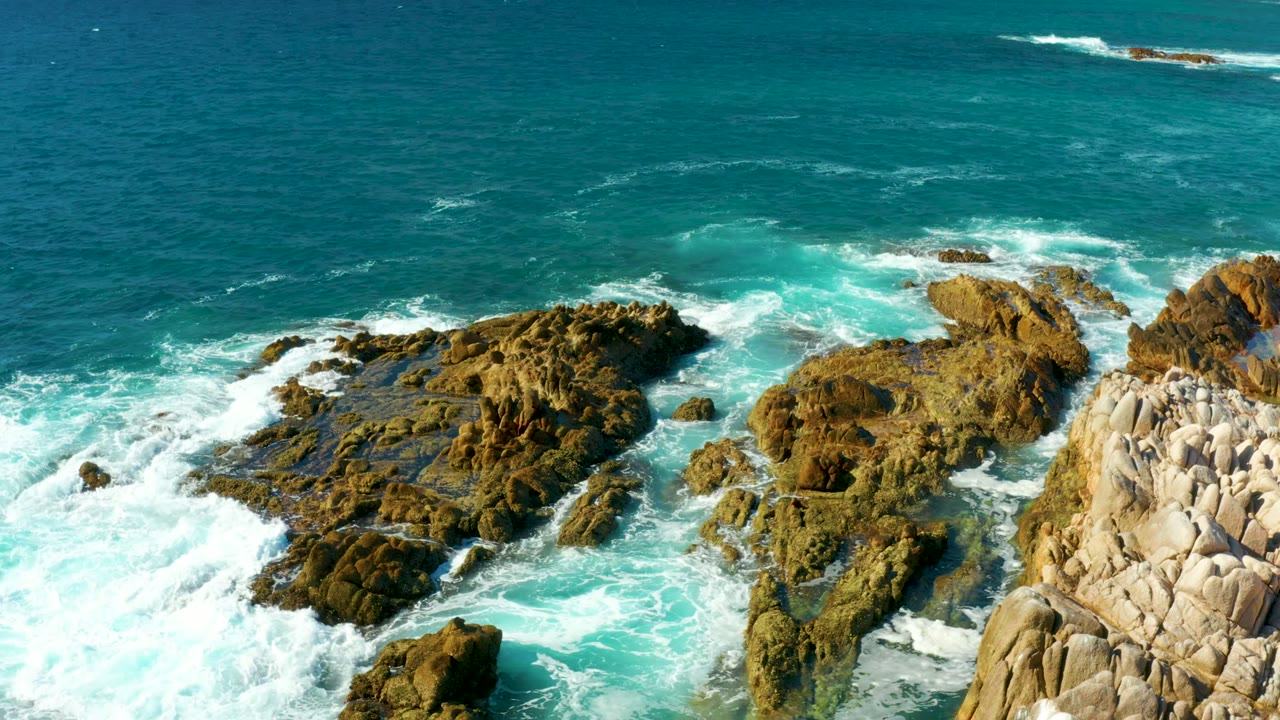 Aerial view of waves curling into brown rocks beside pale coastal formations at Chileno Beach in Los Cabos, with no visible people.