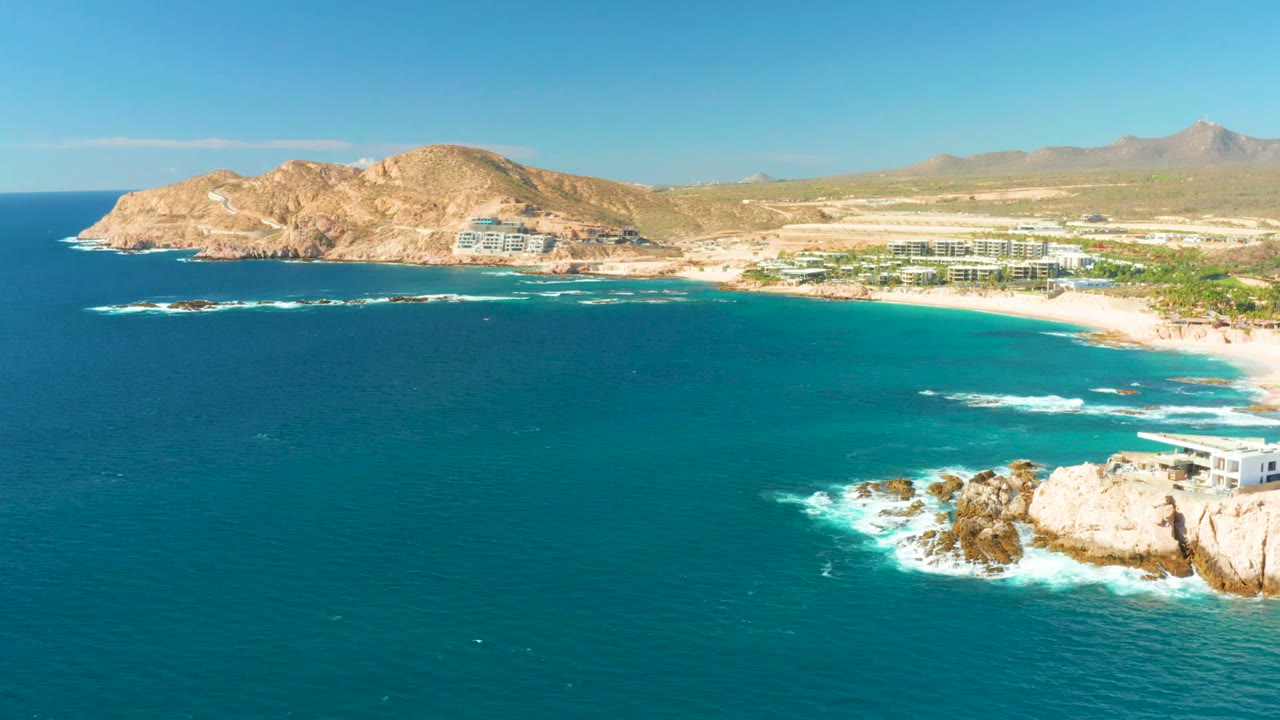 Opening frame of Chileno Beach in Los Cabos with deep blue water in the foreground, turquoise shallows near the sand, rocky shoreline at right, coastal buildings inland, and no visible people.