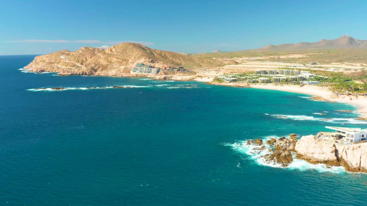 Aerial coastline view with waves breaking near rocks and beach buildings inland.