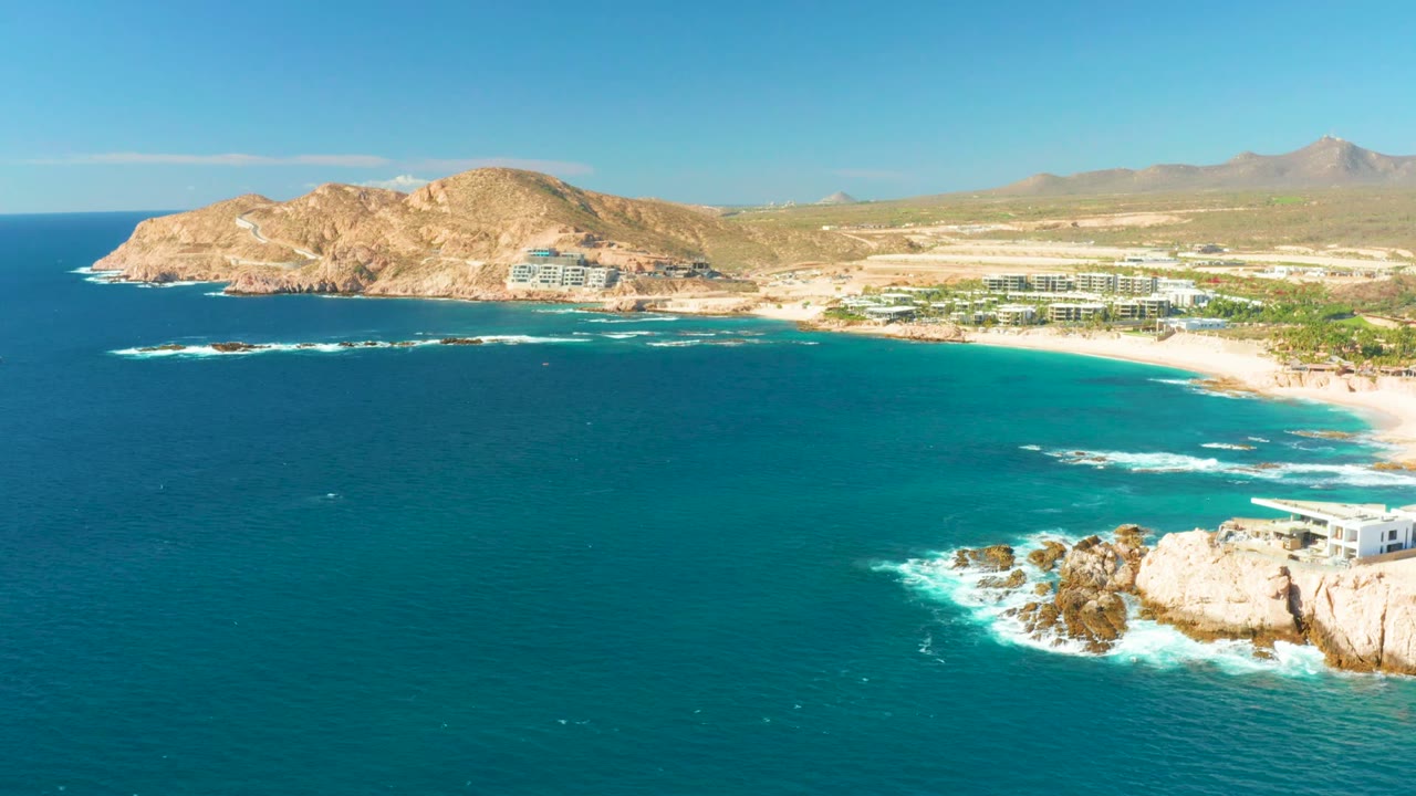 A wide Chileno Beach aerial frame shows the rocky point at right, turquoise water spread across the bay, whitecaps near the rocks, beachfront development, dry hills, and no visible people.