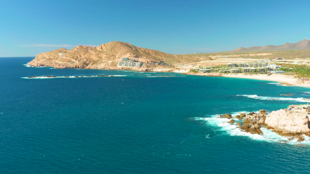 Opening frame over Chileno Beach in Los Cabos shows turquoise water in front, surf hitting rocks at right, beach buildings along the shore, dry hills beyond, and no visible people.