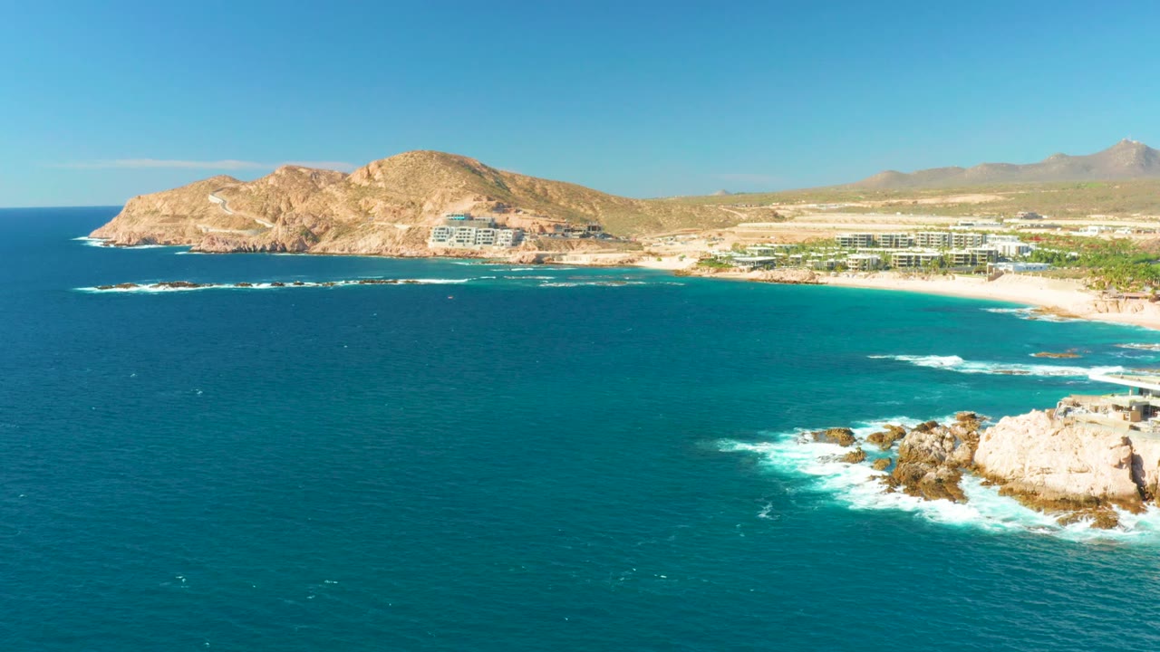 Wide coastal aerial with waves foaming around rocks and beach buildings beyond.