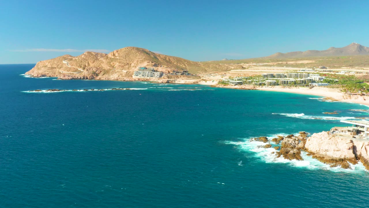At 0:02, the aerial view shows a broad band of turquoise ocean, a foamy break near the rocky point at right, the sandy Chileno Beach shoreline, scattered buildings, dry hills, and no visible people.