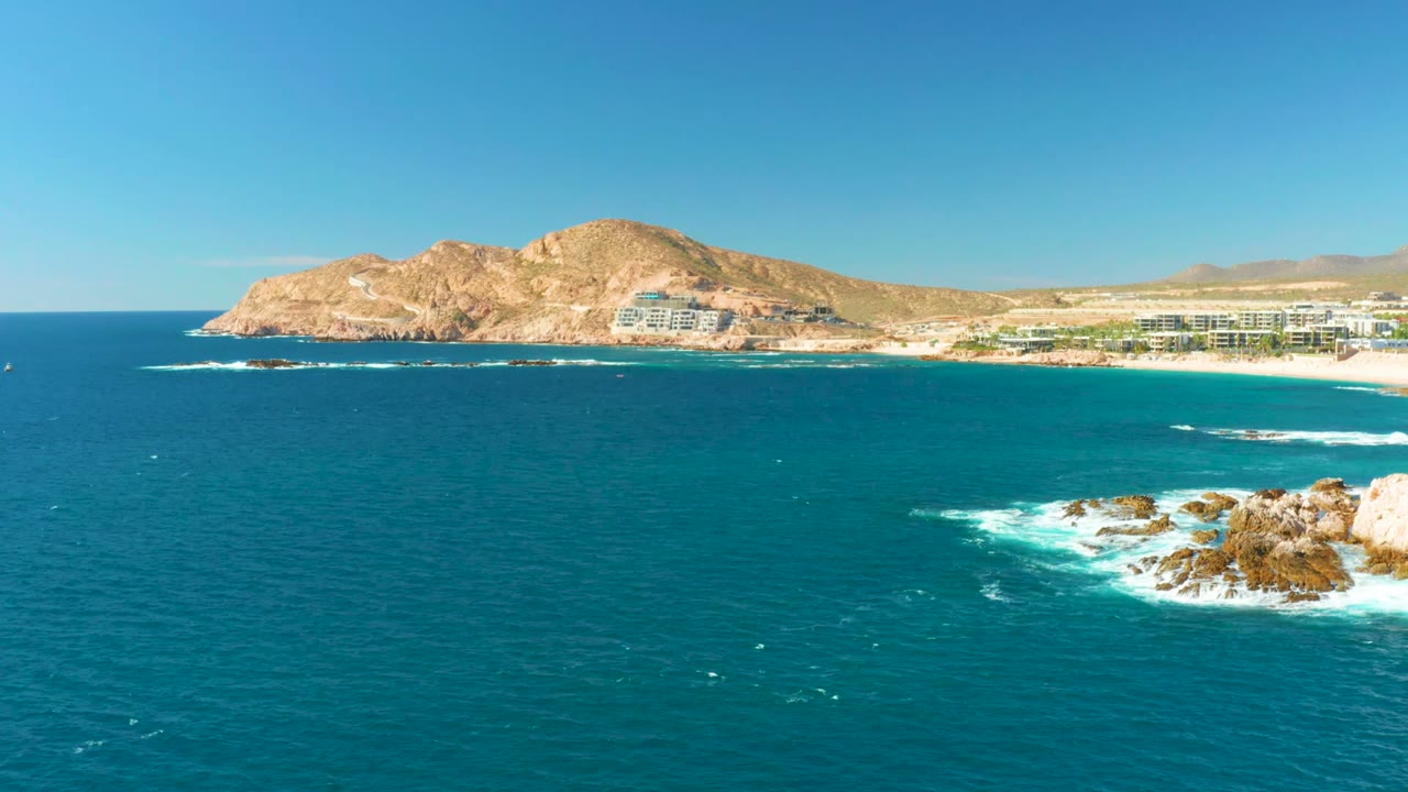 Opening frame of Chileno Beach in Los Cabos, with turquoise water across the foreground, surf on rocks at right, distant beach buildings below tan hills, and no visible people.