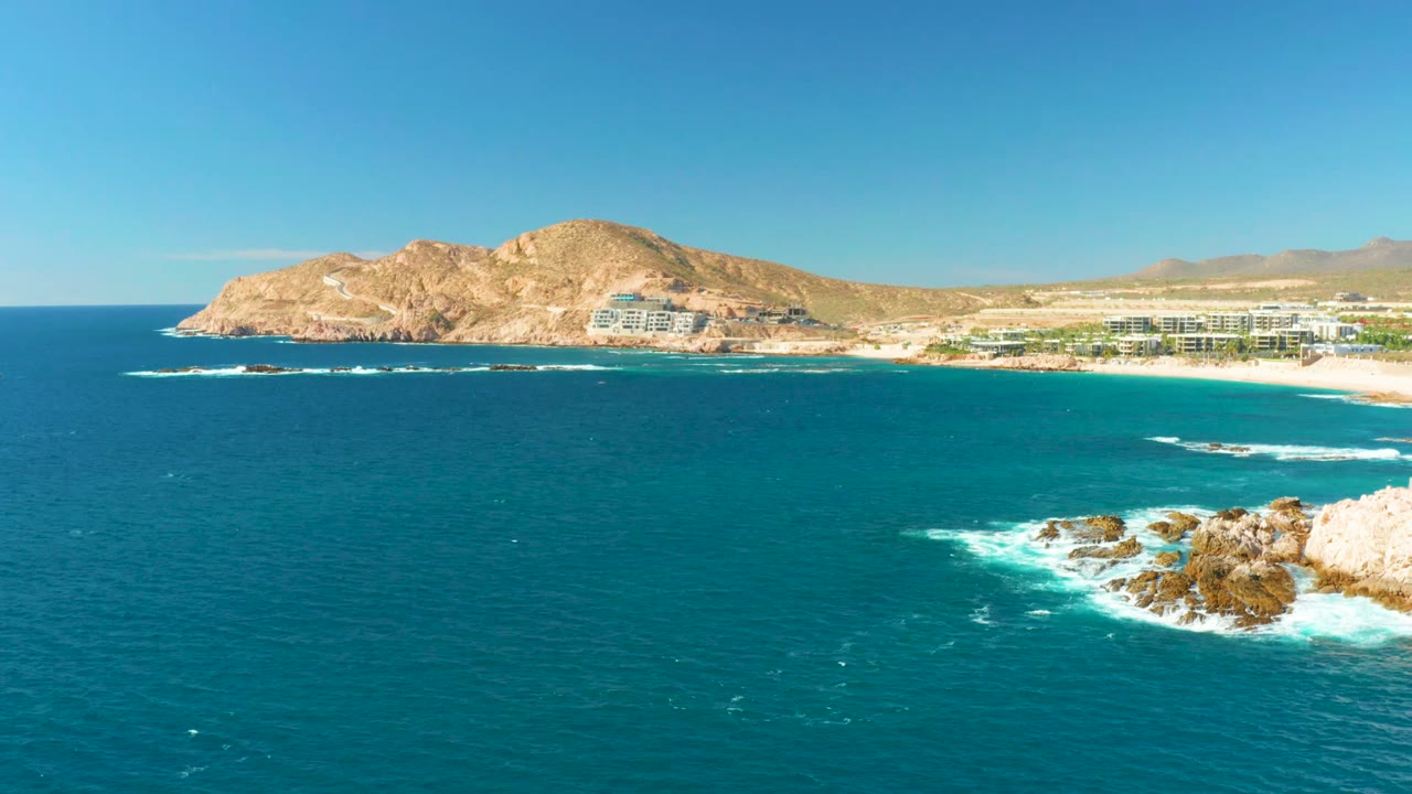 At 0:02, blue-green ocean dominates the frame, the rocky surf sits farther into the lower right, and the Chileno Beach buildings and tan headland stay clear in the distance with no visible people.