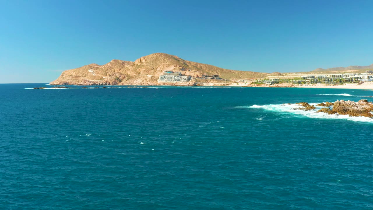 At Chileno Beach in Los Cabos, blue ocean fills the foreground while white surf breaks on rocks at right, with beach buildings and a dry hill beyond, no visible people.