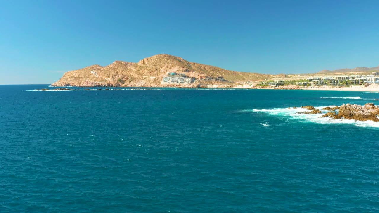 Wide offshore view with teal water, breaking foam on rocks, beach buildings, and hillside.