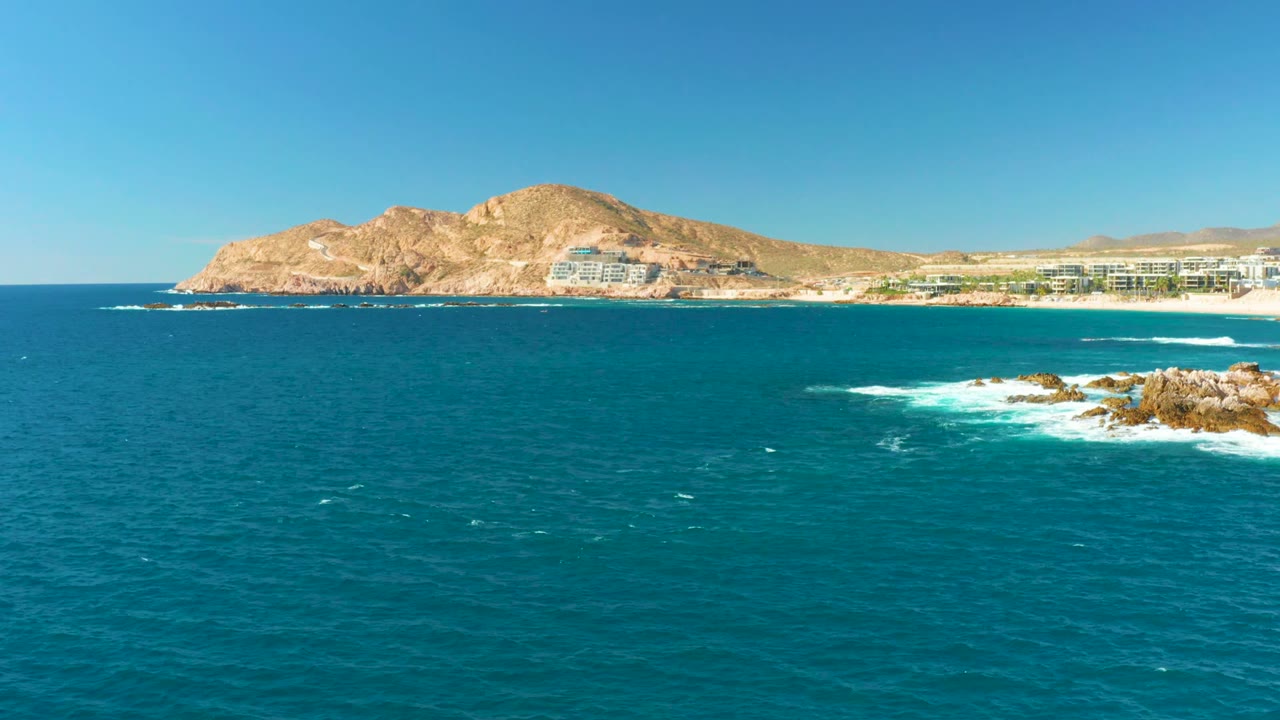 At Chileno Beach in Los Cabos, the right-side rocks sit closer to the lower edge with foam curling around them and low coastal buildings beneath the hill, no visible people.