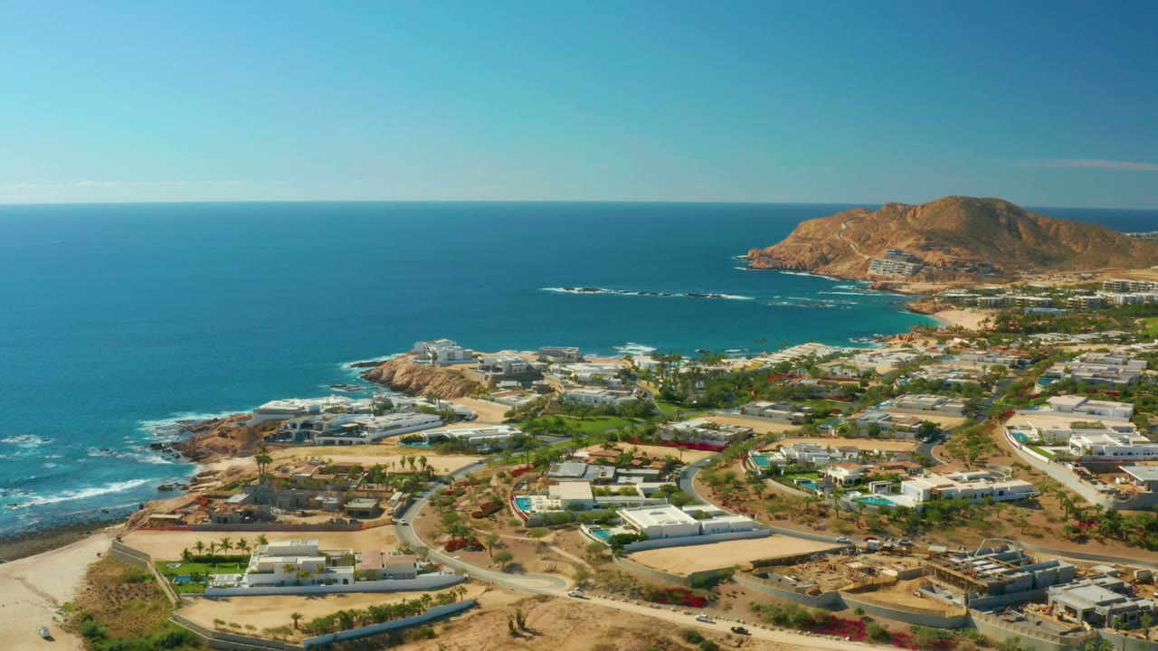 Aerial daytime view over Chileno Beach Los Cabos with coastal homes, winding roads, blue ocean, rocky shoreline, dry hills, and no visible people.