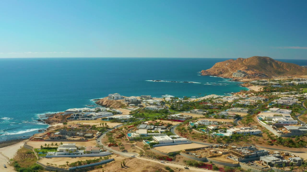 Wide aerial view of Chileno Beach Los Cabos with blue water across the upper frame, surf along the rocky edge, residential roads below, and no visible people.