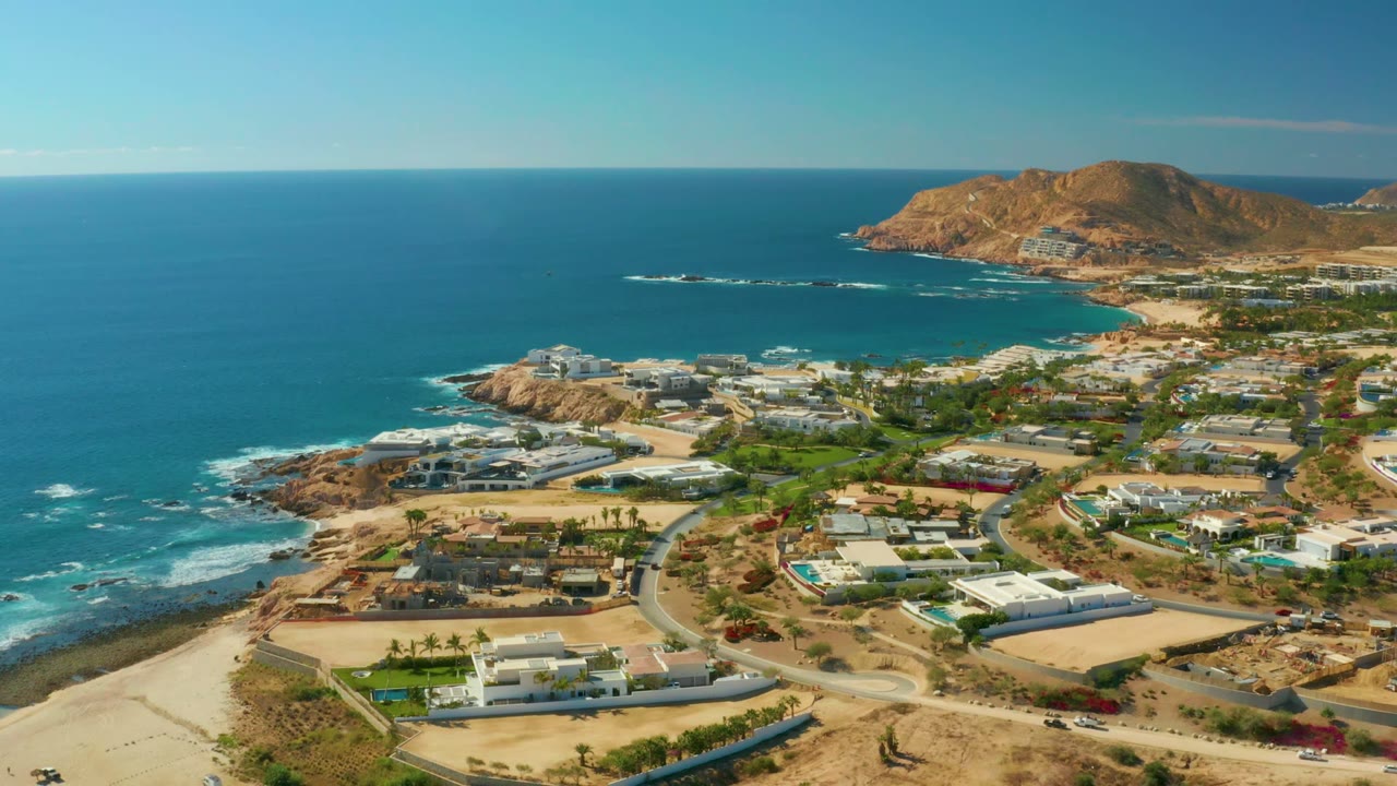 Wide aerial over Chileno Beach Los Cabos with resort buildings and roads in the foreground, turquoise ocean at left, rocky points, distant hills, and no visible people.