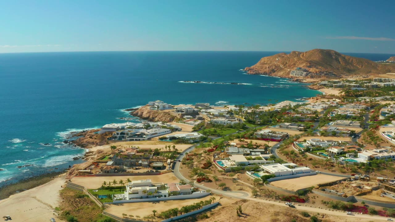 Aerial frame of Chileno Beach Los Cabos showing more open ocean on the left, low white buildings near the shore, curving roads inland, a brown hill in the background, and no visible people.