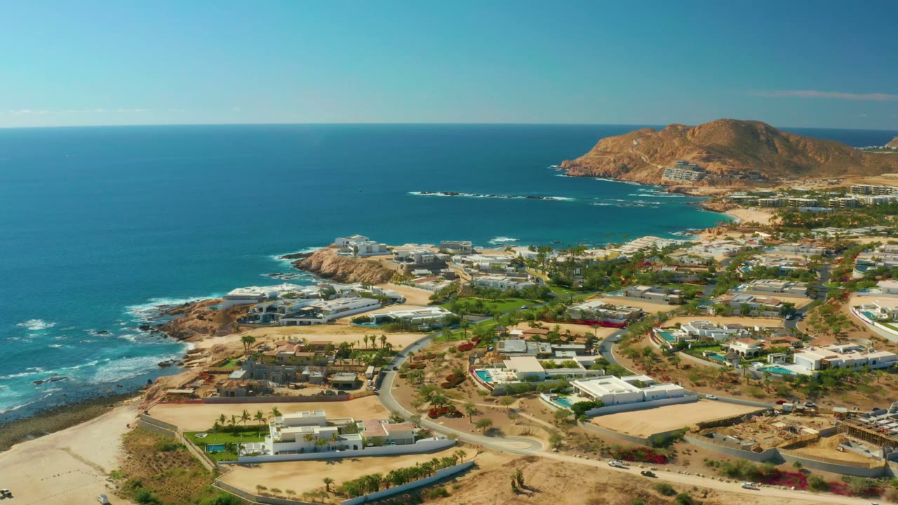 Aerial view over Chileno Beach Los Cabos with blue water across the left half, resort buildings and landscaped lots on the right, small surf lines on offshore rocks, and no visible people.