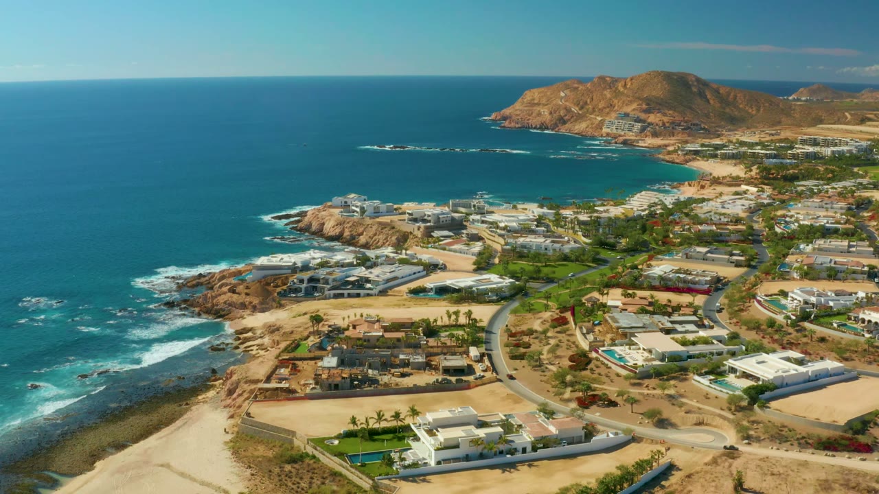 Aerial frame over Chileno Beach, Los Cabos, showing blue ocean at left, surf along the rocky shoreline, beach homes and roads below, brown hills in the distance, and no visible people.